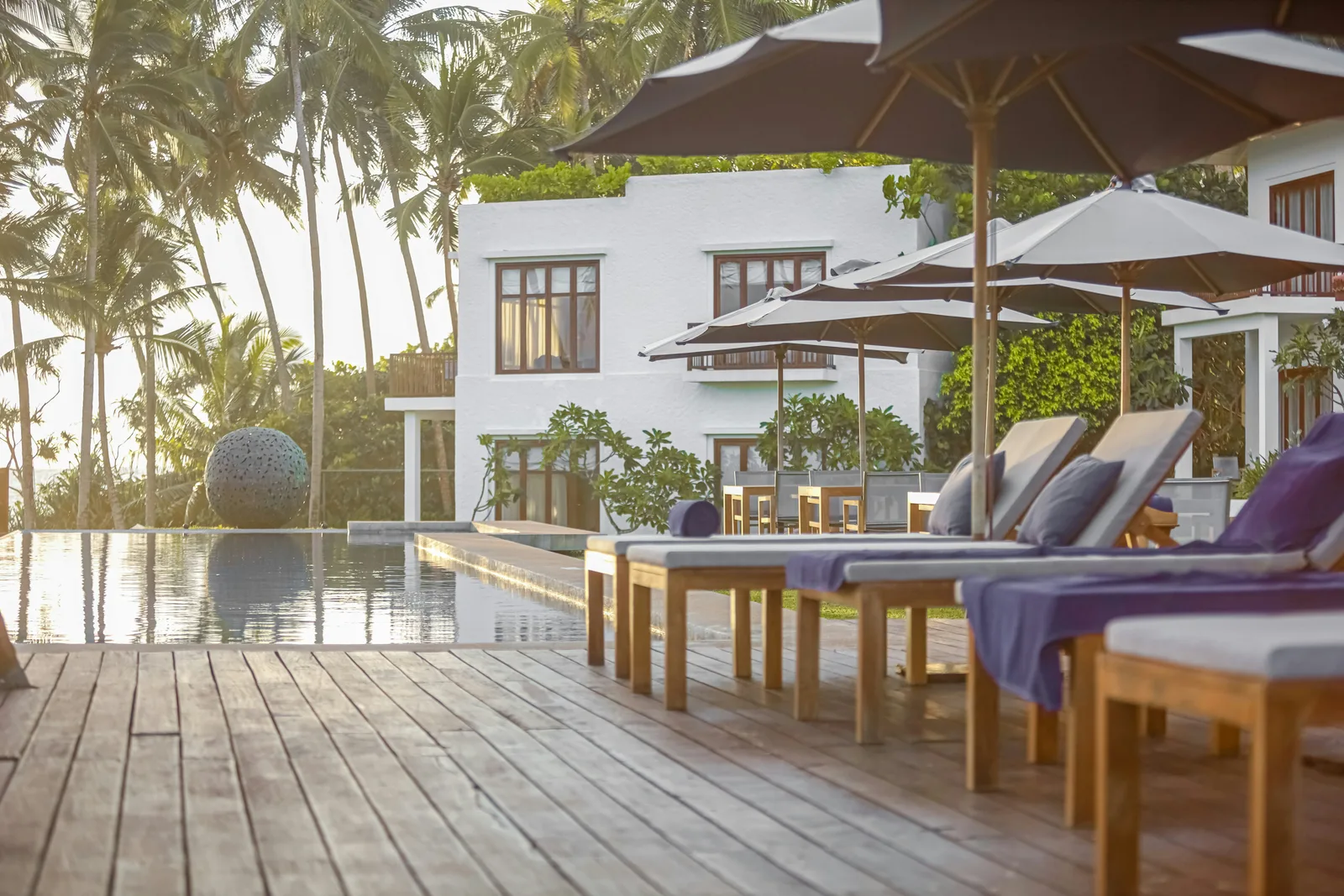 Empty tropical pool patio with wood decking, lounge chairs, umbrellas, and a paved pool edge