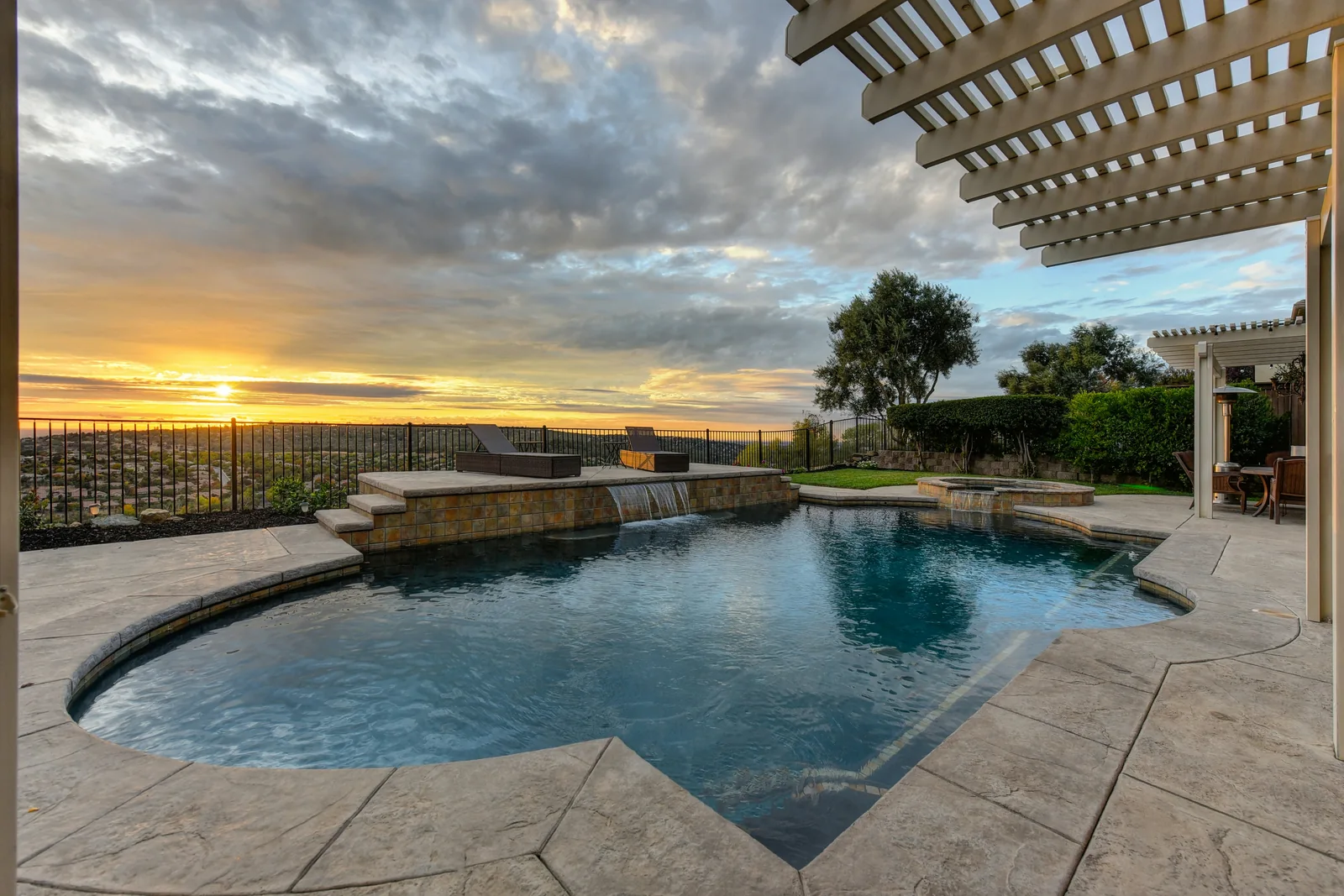 Empty backyard pool with pergola beams, patio paving, and a perimeter fence at sunset